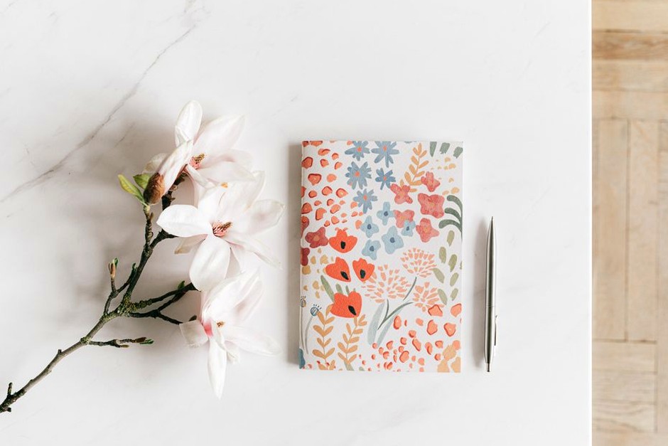 A floral notebook, pen and magnolia branch arranged on a bright desk.